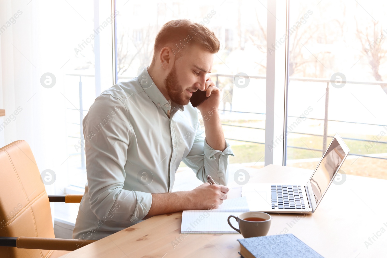 Photo of Young man talking on mobile phone while working with laptop at desk. Home office