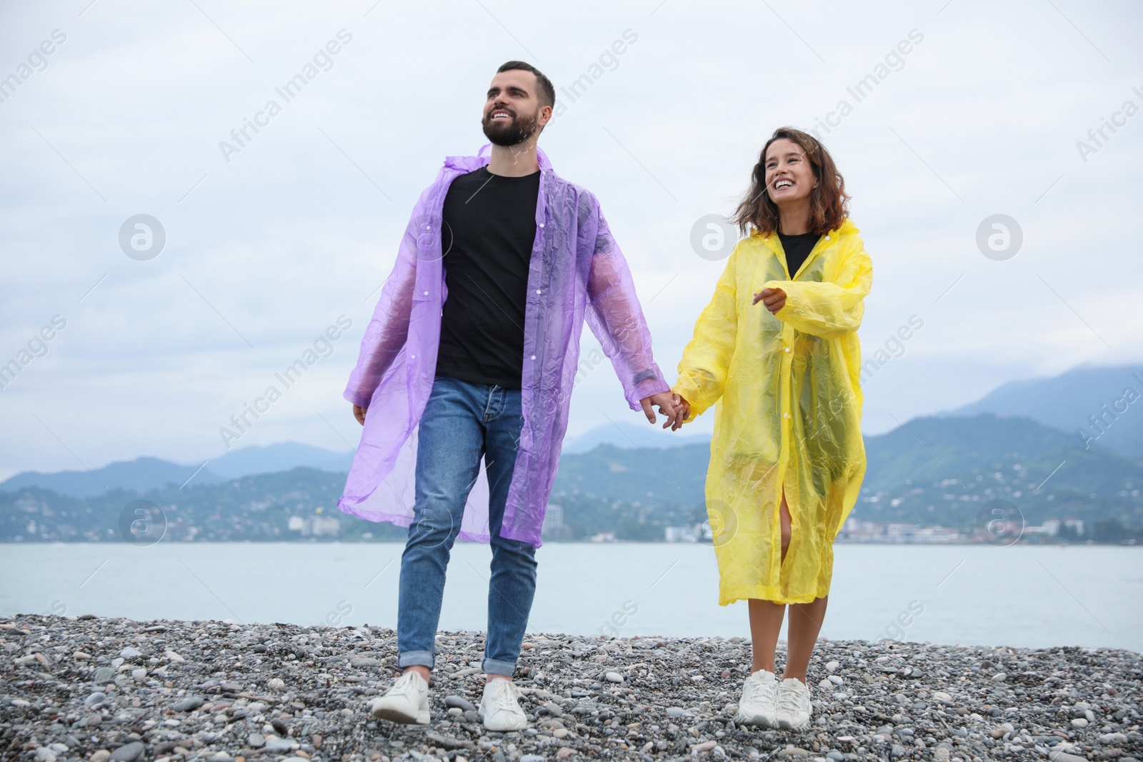 Photo of Young couple in raincoats enjoying time together under rain on beach