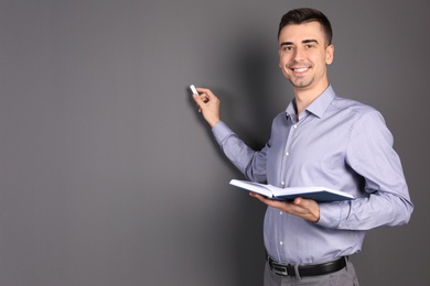 Young male teacher with book and chalk on grey background