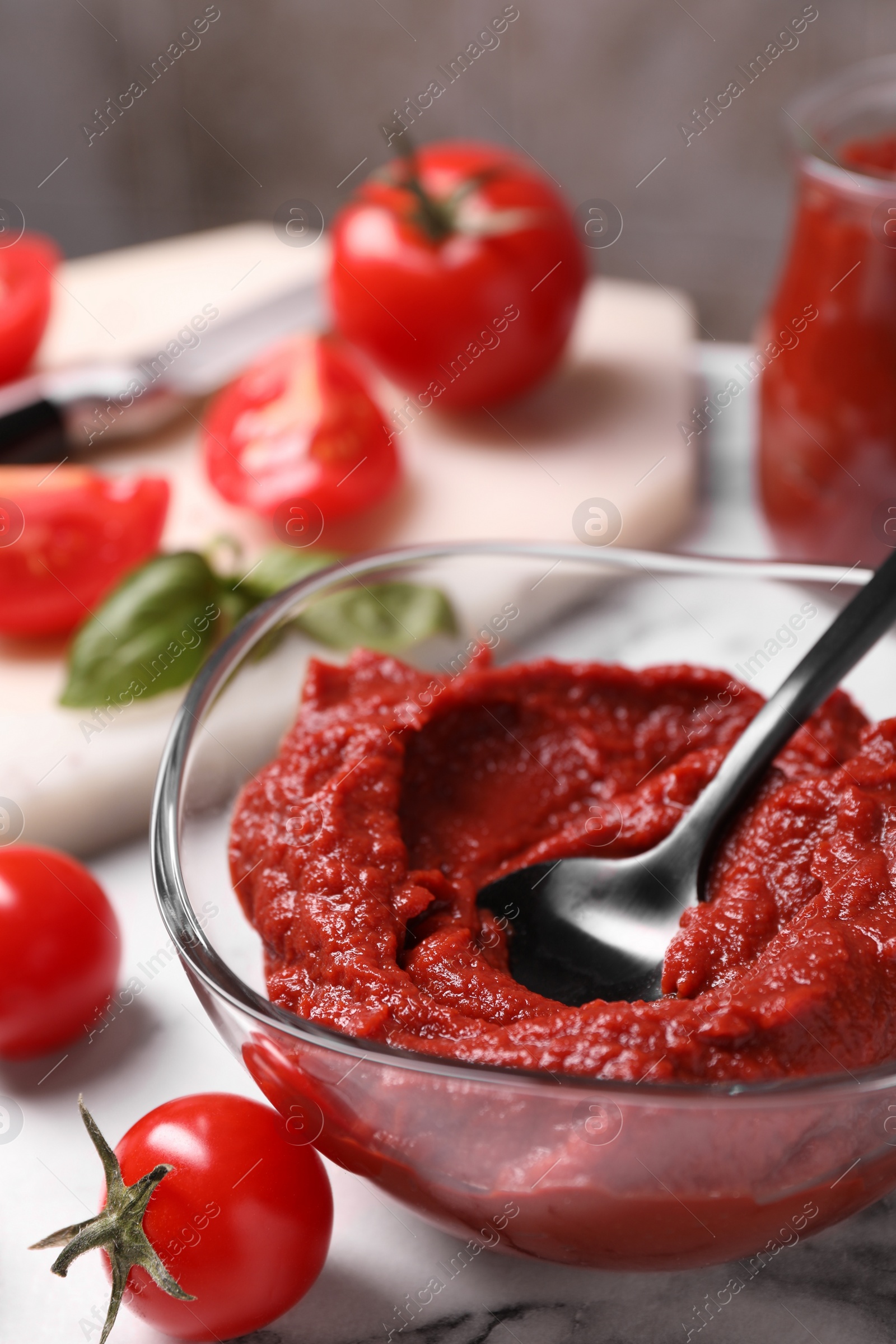 Photo of Glass bowl of tasty tomato paste with spoon and ingredients on white marble table, closeup