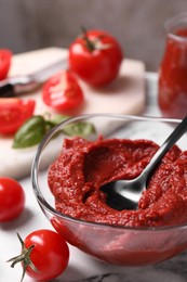 Glass bowl of tasty tomato paste with spoon and ingredients on white marble table, closeup