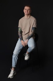 Photo of Smiling young man with tattoos sitting on stool against black background