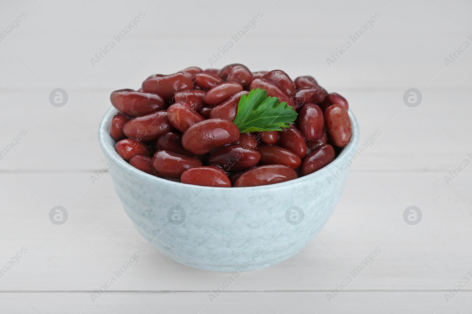 Photo of Bowl of canned kidney beans with parsley on white wooden table, closeup