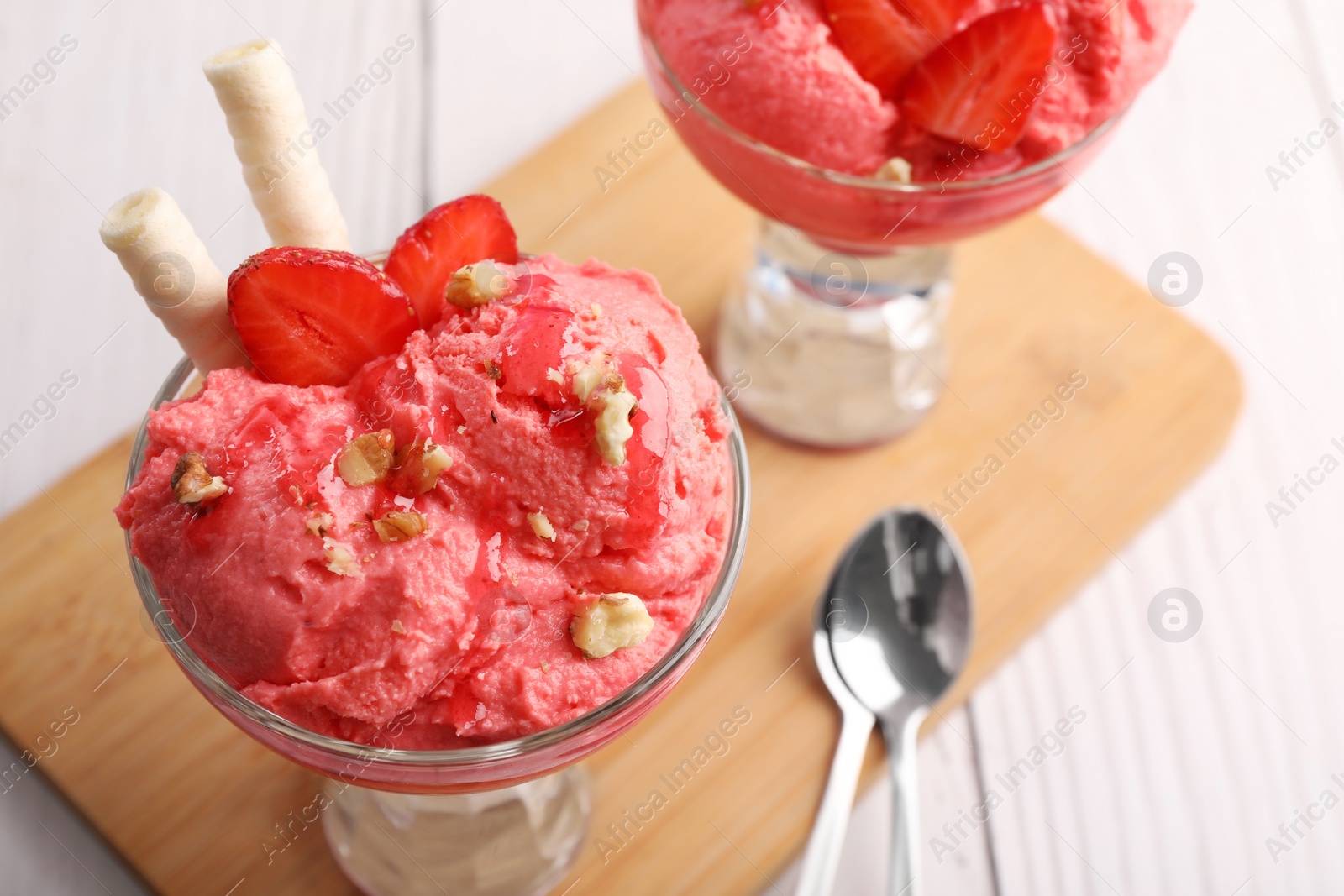 Photo of Delicious scoops of strawberry ice cream with wafer sticks and nuts in glass dessert bowls served on white wooden table, closeup