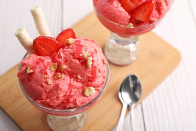 Delicious scoops of strawberry ice cream with wafer sticks and nuts in glass dessert bowls served on white wooden table, closeup