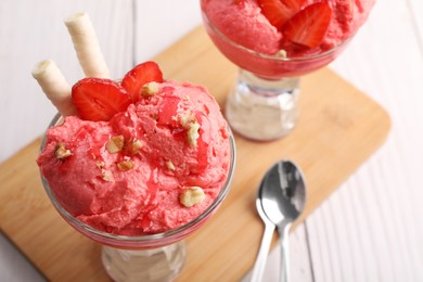 Photo of Delicious scoops of strawberry ice cream with wafer sticks and nuts in glass dessert bowls served on white wooden table, closeup