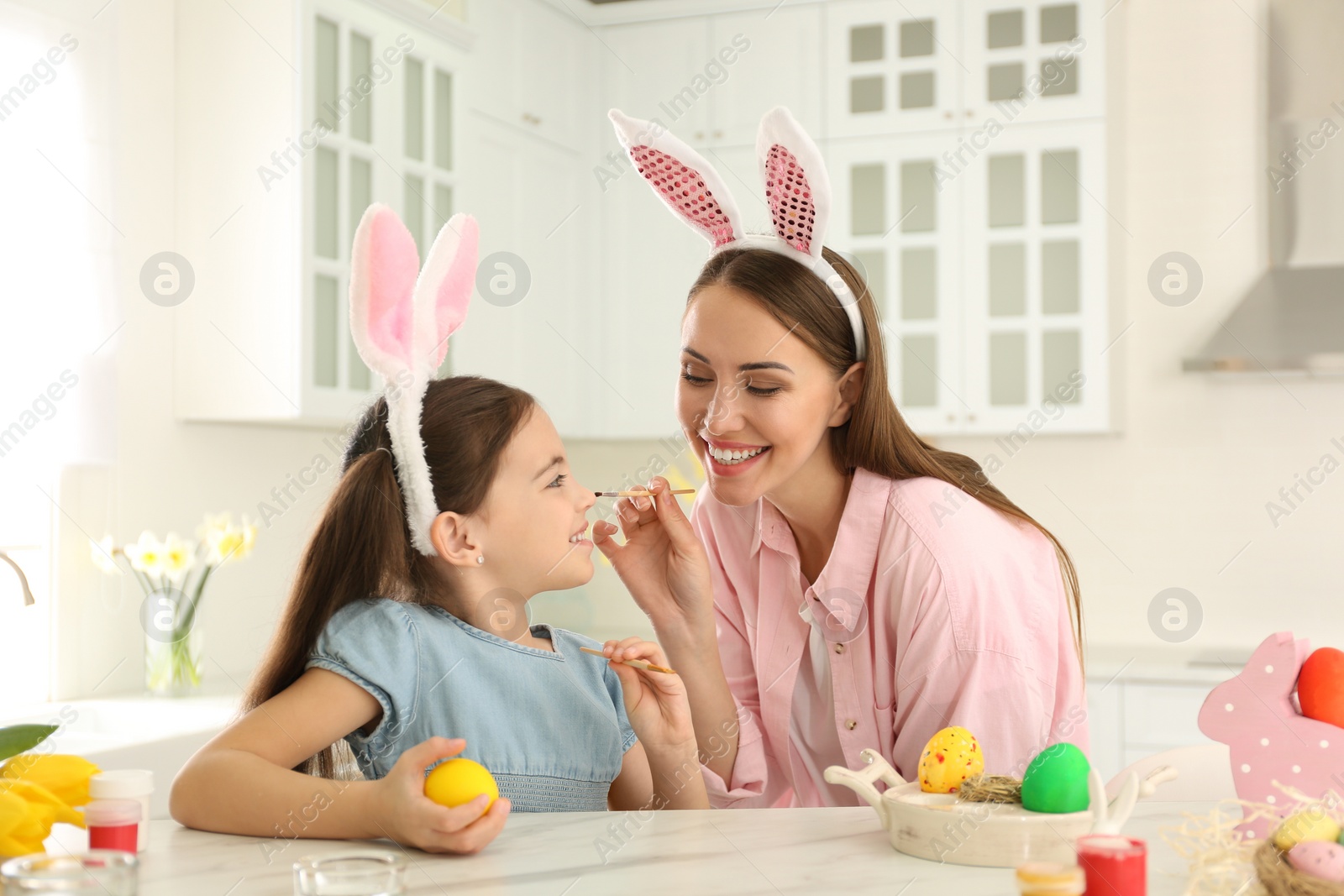 Photo of Happy mother and daughter with bunny ears headbands having fun while painting Easter egg in kitchen