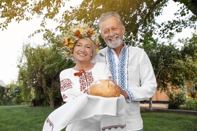Photo of Happy mature couple in national clothes with bread and salt outdoors. Ukrainian welcoming tradition