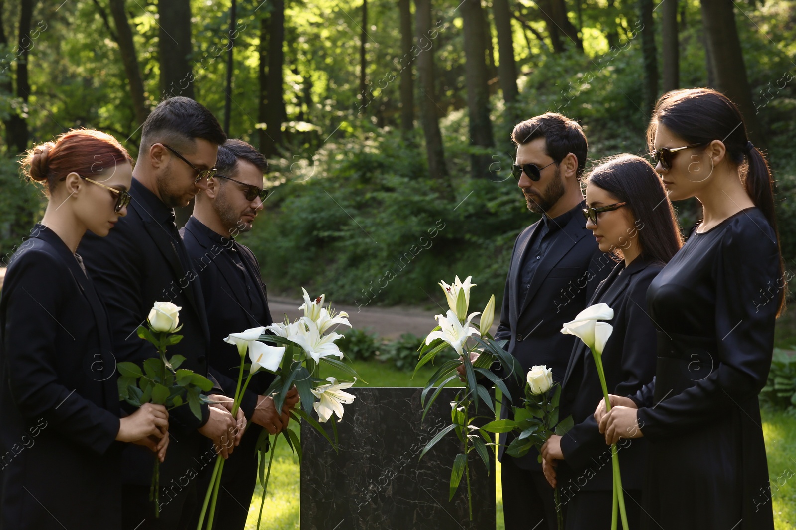 Photo of Sad people with flowers mourning near granite tombstone at cemetery outdoors. Funeral ceremony