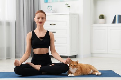 Beautiful woman with cute red cat practicing yoga on mat at home