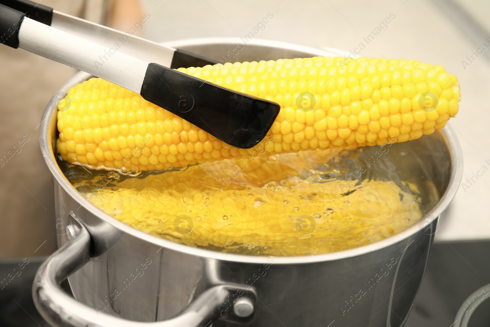 Photo of Taking boiled corn from pot with tongs in kitchen, closeup