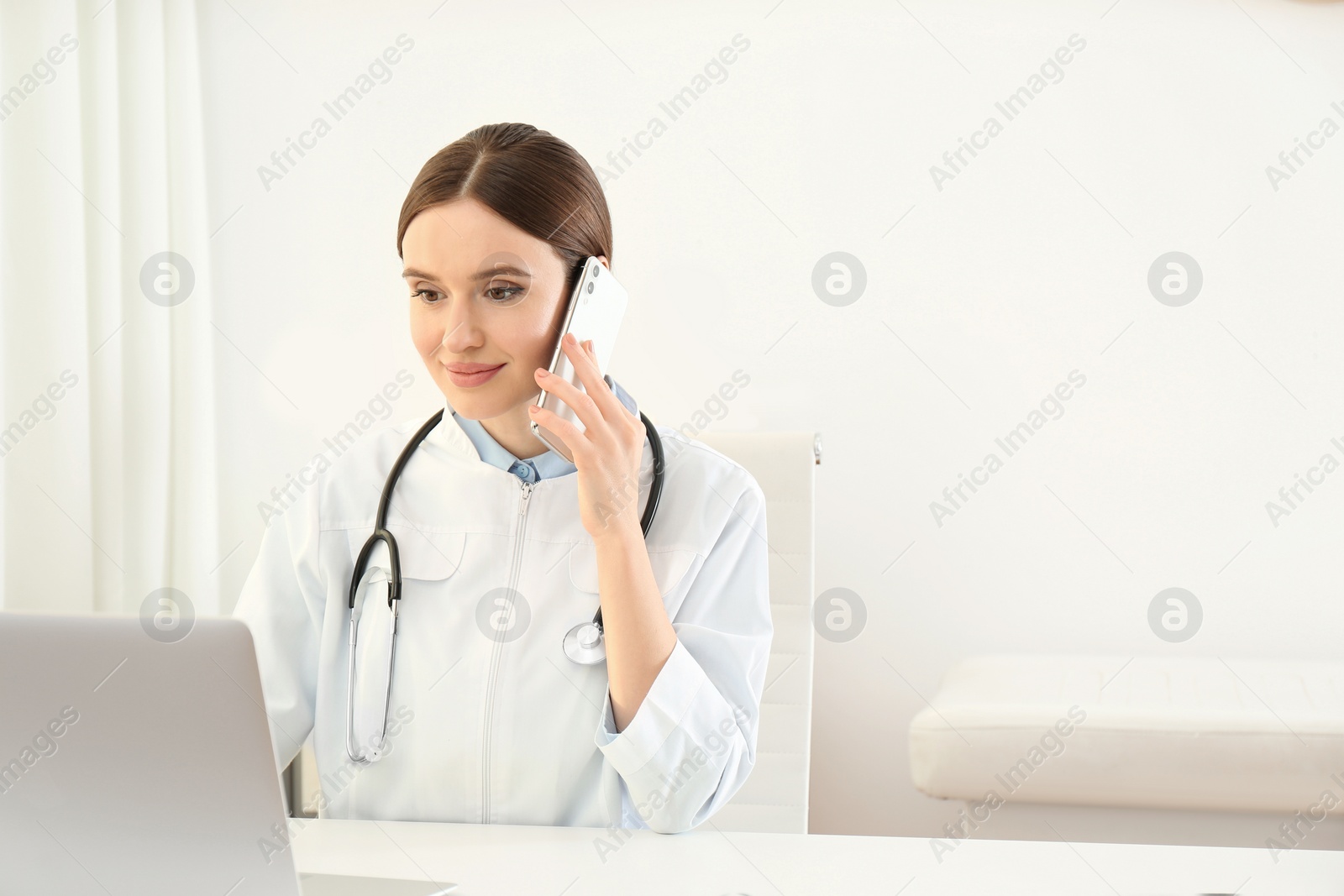 Photo of Young female doctor talking on phone at table in office