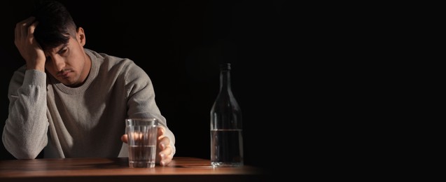 Image of Suffering from hangover. Man with alcoholic drink at table against black background, space for text. Banner design