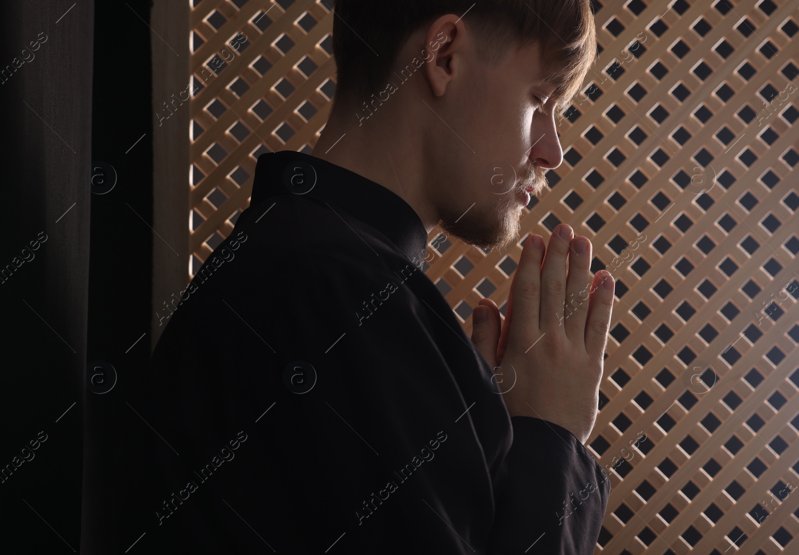 Photo of Catholic priest praying near wooden window in confessional booth