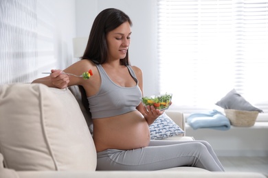 Photo of Young pregnant woman with bowl of vegetable salad in living room. Taking care of baby health