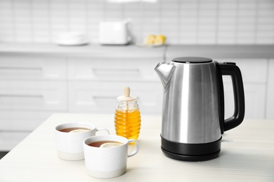 Modern electric kettle, cups of tea and honey on wooden table in kitchen