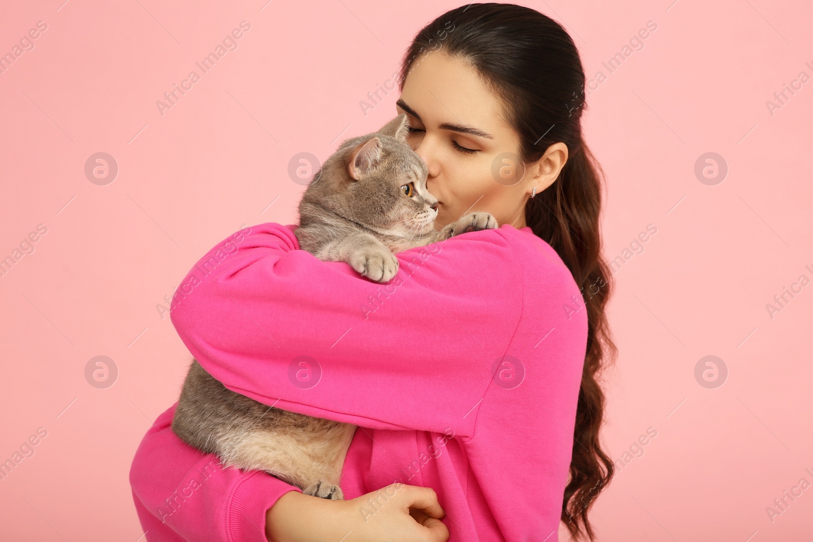 Photo of Young woman kissing her adorable cat on pink background