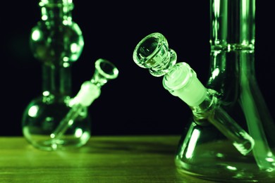 Photo of Glass bongs on wooden table against black background, toned in green. Smoking device