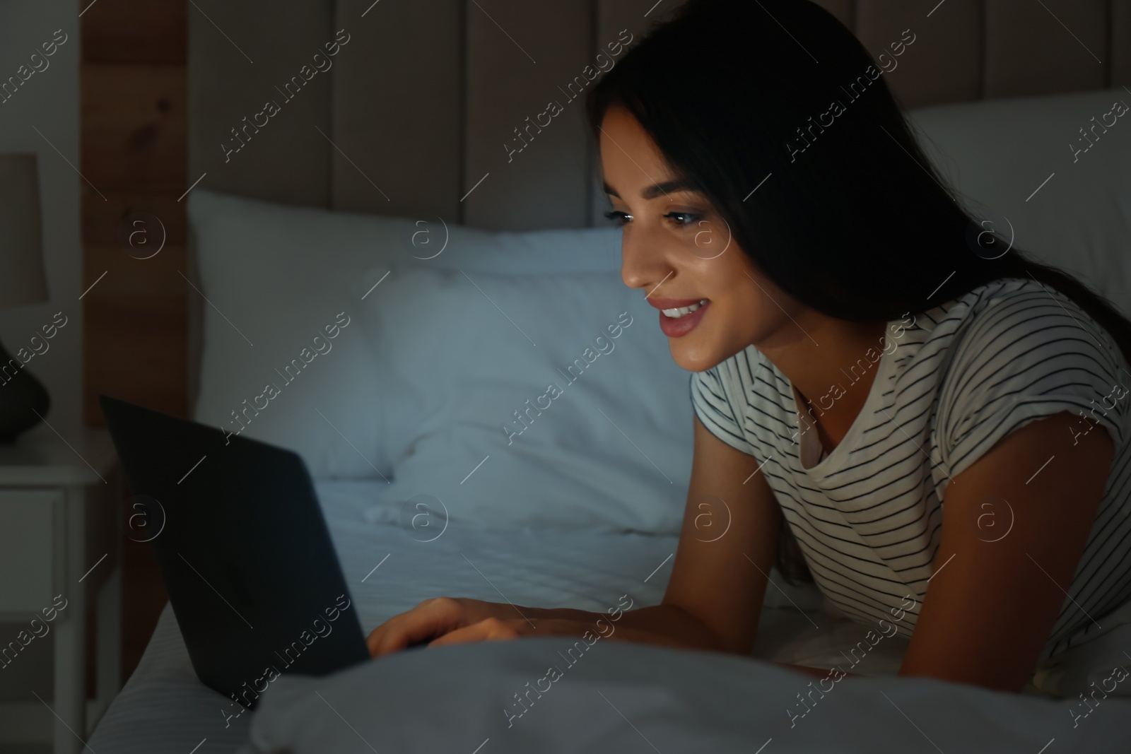 Photo of Happy woman using laptop in dark bedroom