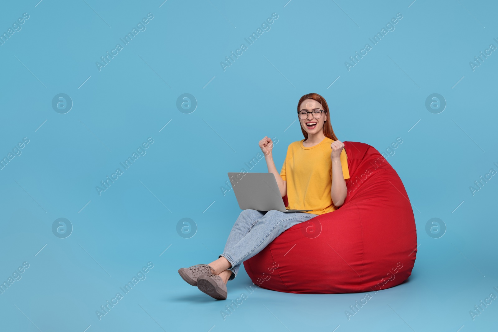 Photo of Happy young woman with laptop sitting on beanbag chair against light blue background, space for text