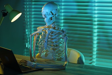 Waiting concept. Human skeleton sitting at wooden table with laptop indoors