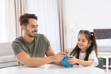 Photo of Little girl with her father putting money into piggy bank at table indoors