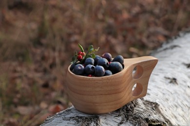 Wooden mug full of fresh ripe blueberries and lingonberries on log in forest