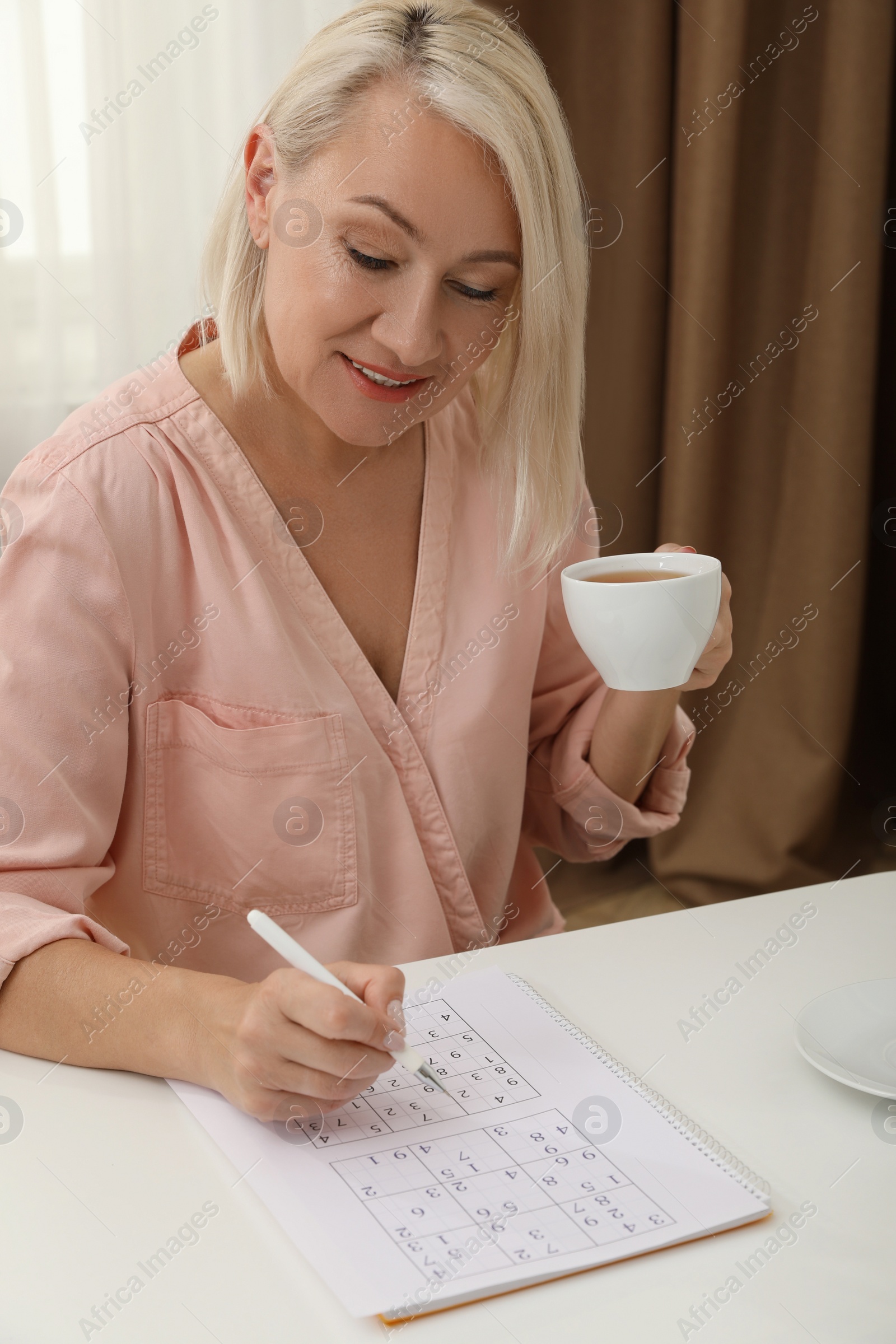 Photo of Middle aged woman with cup of drink solving sudoku puzzle at table indoors