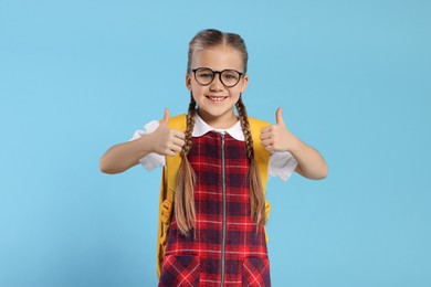 Photo of Happy schoolgirl in glasses with backpack showing thumbs up gesture on light blue background