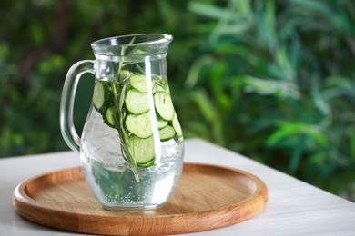 Photo of Refreshing cucumber water with rosemary in jug on white wooden table against blurred green background. Space for text