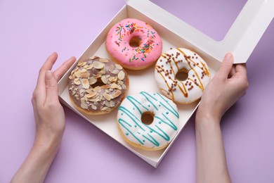 Woman holding box with tasty glazed donuts on violet background, top view