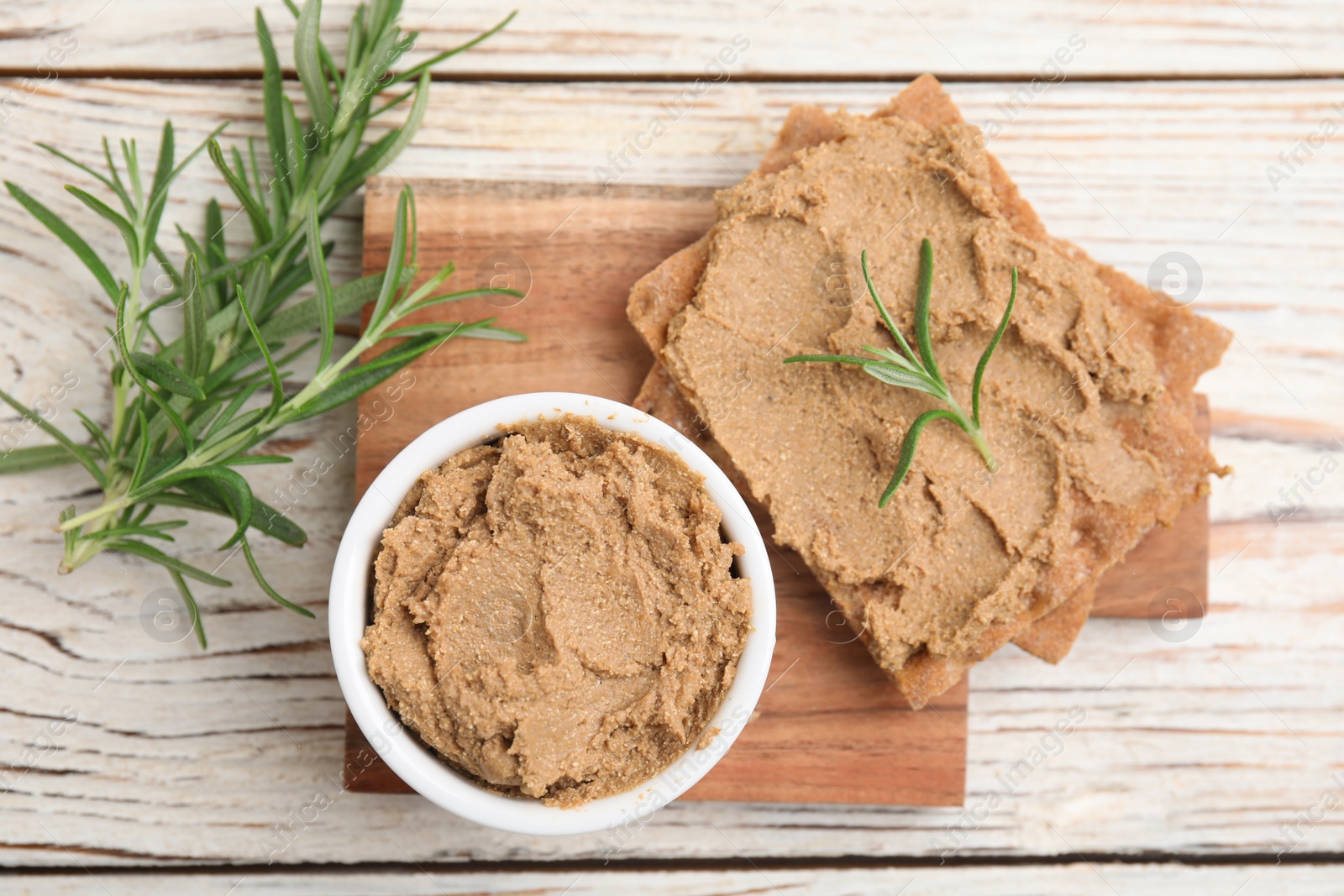 Photo of Crispy crackers with delicious meat pate and rosemary on white wooden table, flat lay