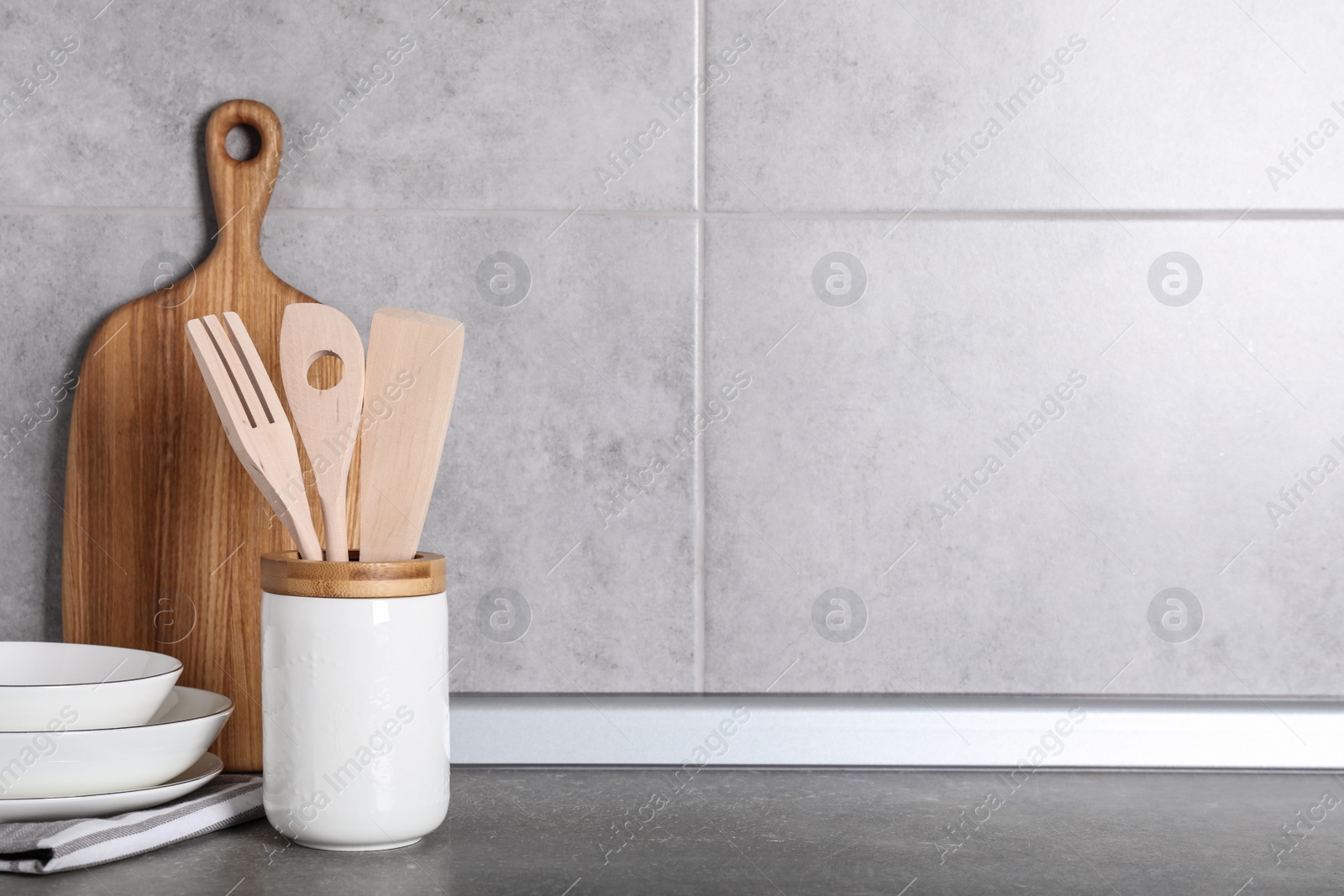 Photo of Holder with kitchen utensils, ceramic dishes and cutting board on grey table. Space for text