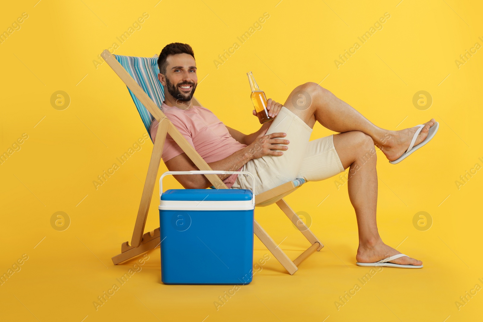 Photo of Happy man with bottle of beer resting in deck chair near cool box on yellow background