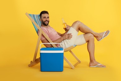 Photo of Happy man with bottle of beer resting in deck chair near cool box on yellow background