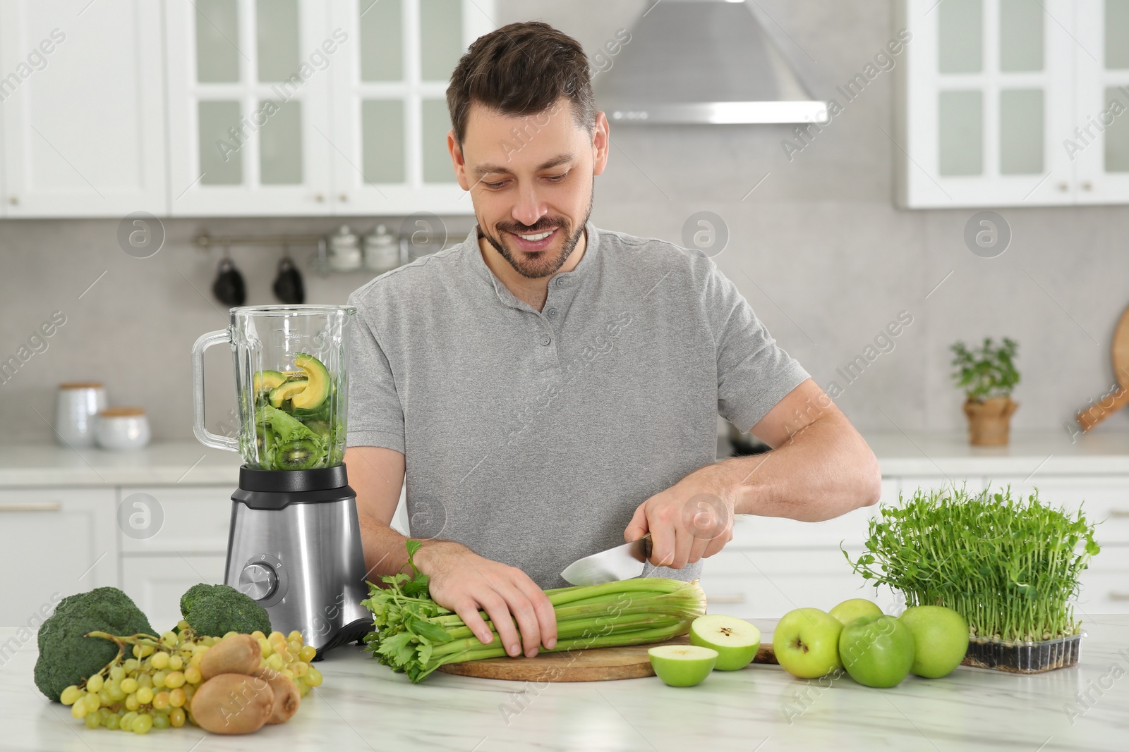 Photo of Happy man cutting celery for delicious smoothie at white marble table in kitchen