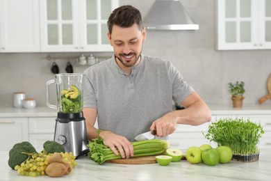 Happy man cutting celery for delicious smoothie at white marble table in kitchen