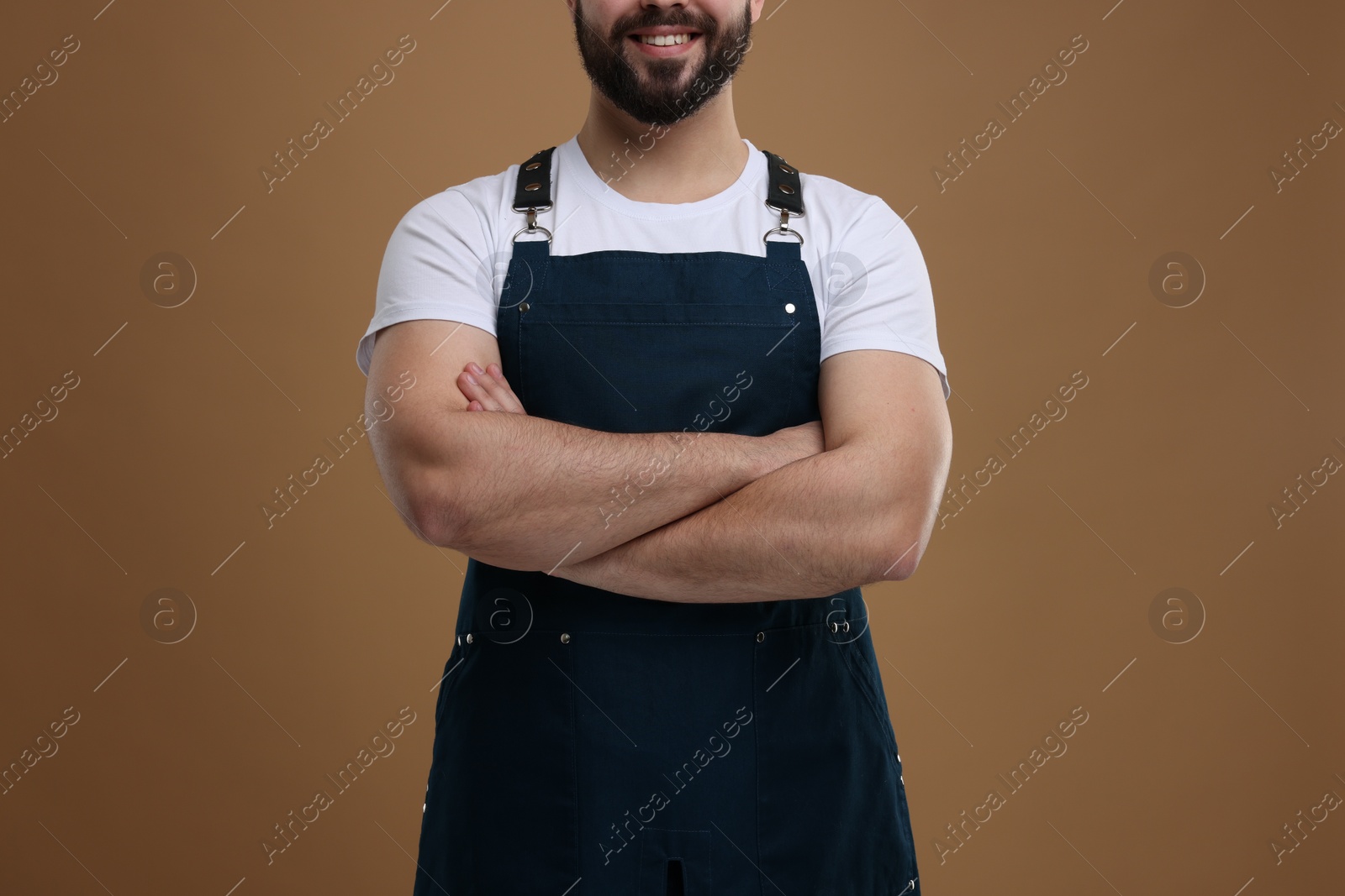 Photo of Smiling man in kitchen apron with crossed arms on brown background, closeup. Mockup for design