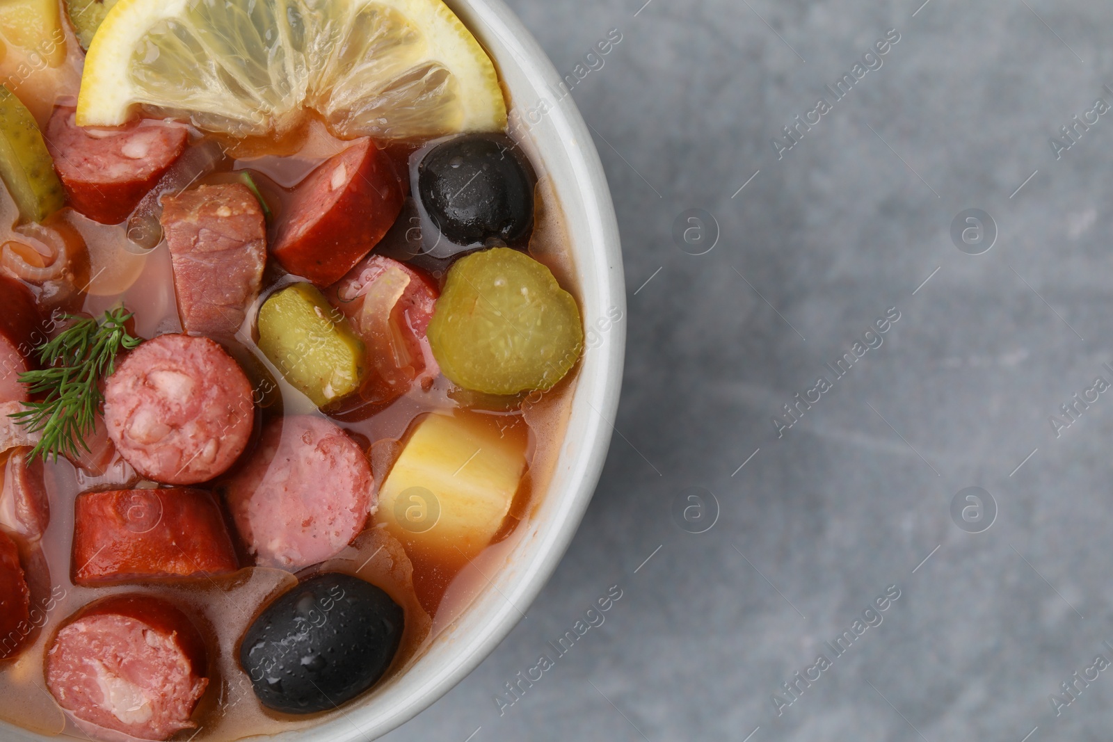 Photo of Meat solyanka soup with thin dry smoked sausages in bowl on grey table, top view. Space for text