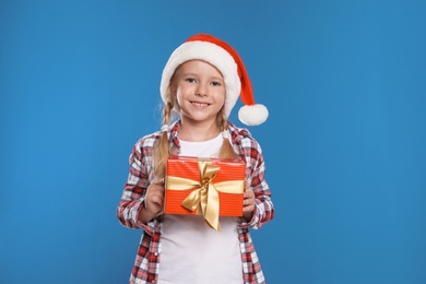 Photo of Happy little child in Santa hat with gift box on blue background. Christmas celebration