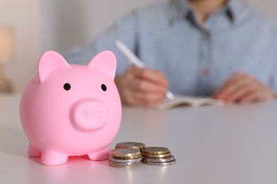 Photo of Woman at white table, focus on pink piggy bank. Space for text