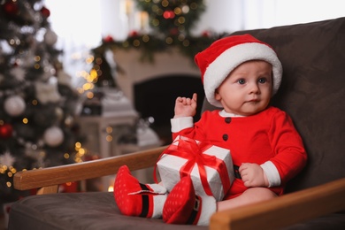 Cute little baby wearing Santa Claus suit with Christmas gift in armchair at home