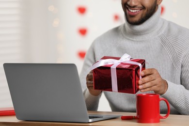 Photo of Valentine's day celebration in long distance relationship. Man holding gift box while having video chat with his girlfriend via laptop, closeup