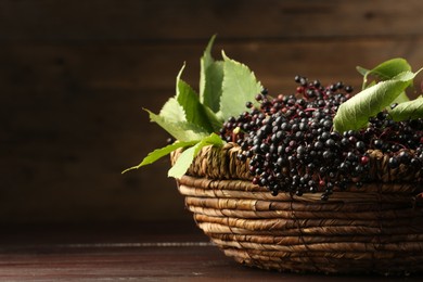 Ripe elderberries with green leaves in wicker basket on wooden table, space for text