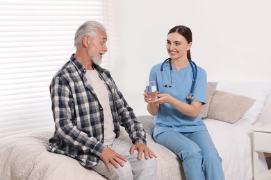 Young healthcare worker giving glass of water to senior man indoors