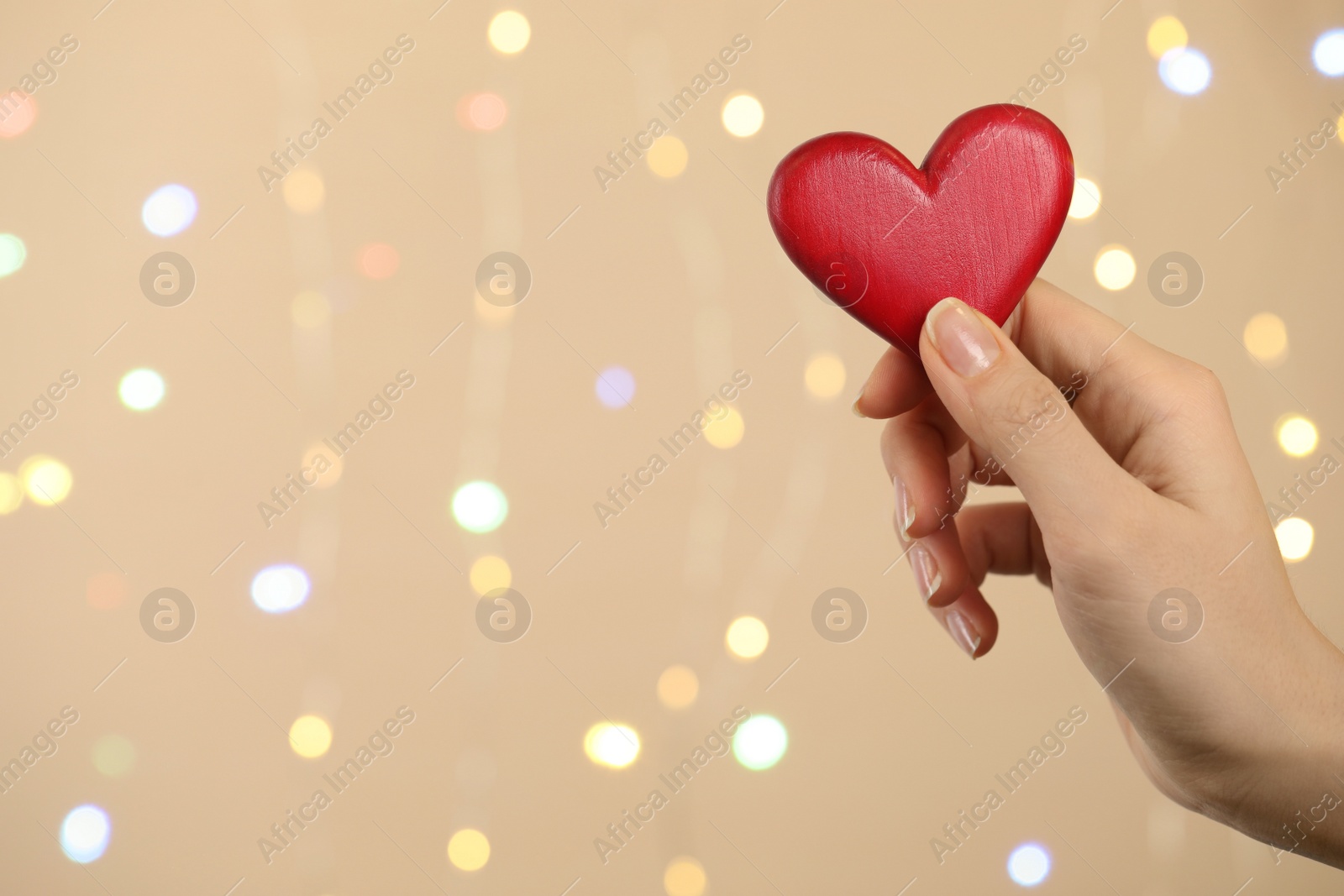 Photo of Closeup view of woman holding red heart on beige background with festive lights, space for text. St. Valentine's day