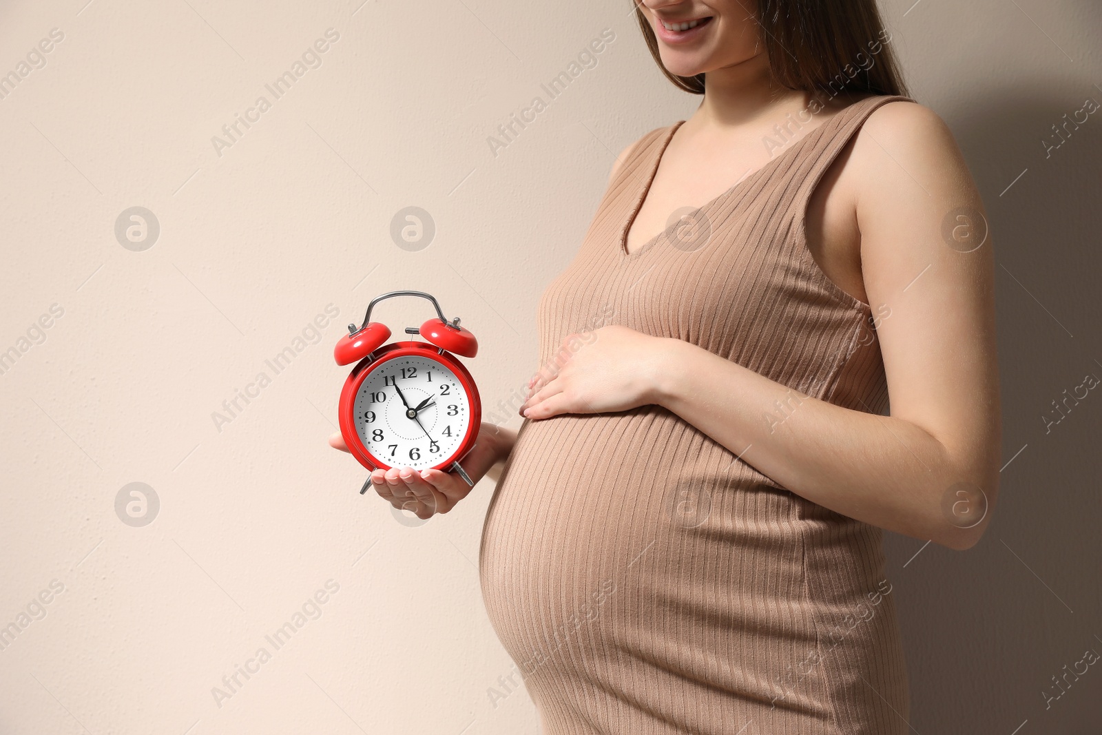 Photo of Young pregnant woman holding alarm clock near her belly on beige background, closeup and space for text. Time to give birth