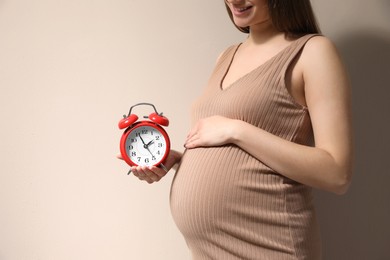 Photo of Young pregnant woman holding alarm clock near her belly on beige background, closeup and space for text. Time to give birth