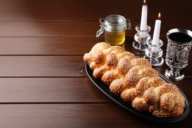 Photo of Homemade braided bread with sesame seeds, goblet, honey and candles on wooden table, space for text. Traditional Shabbat challah
