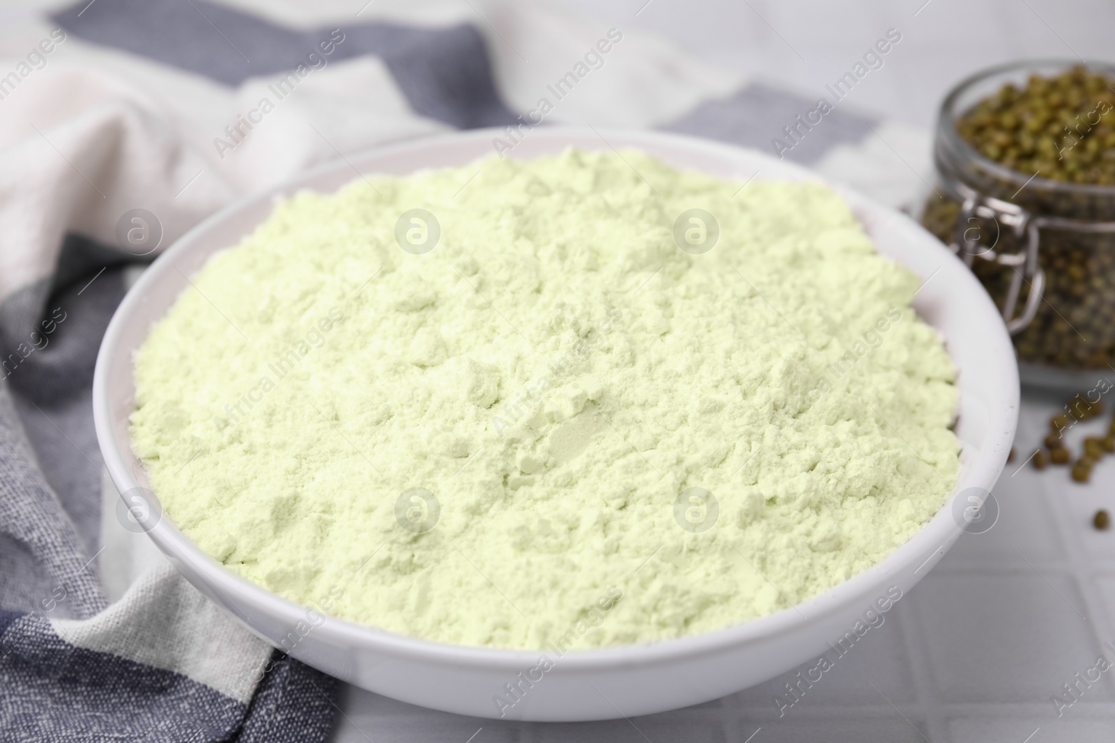 Photo of Mung bean flour in bowl and seeds on white tiled table, closeup